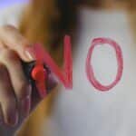 Close-up of teenager writing 'No!' in red on glass, symbolizing refusal, captured in vibrant studio setting.