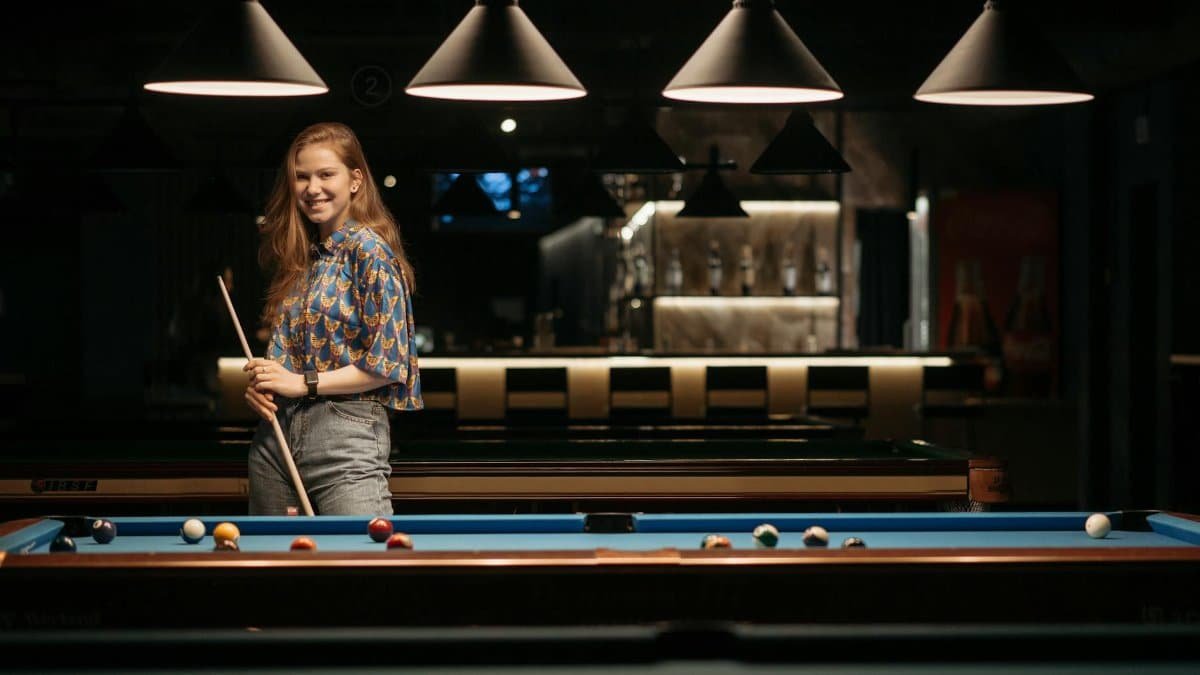 Smiling woman standing by a billiard table holding a cue stick in a dimly lit pool hall.