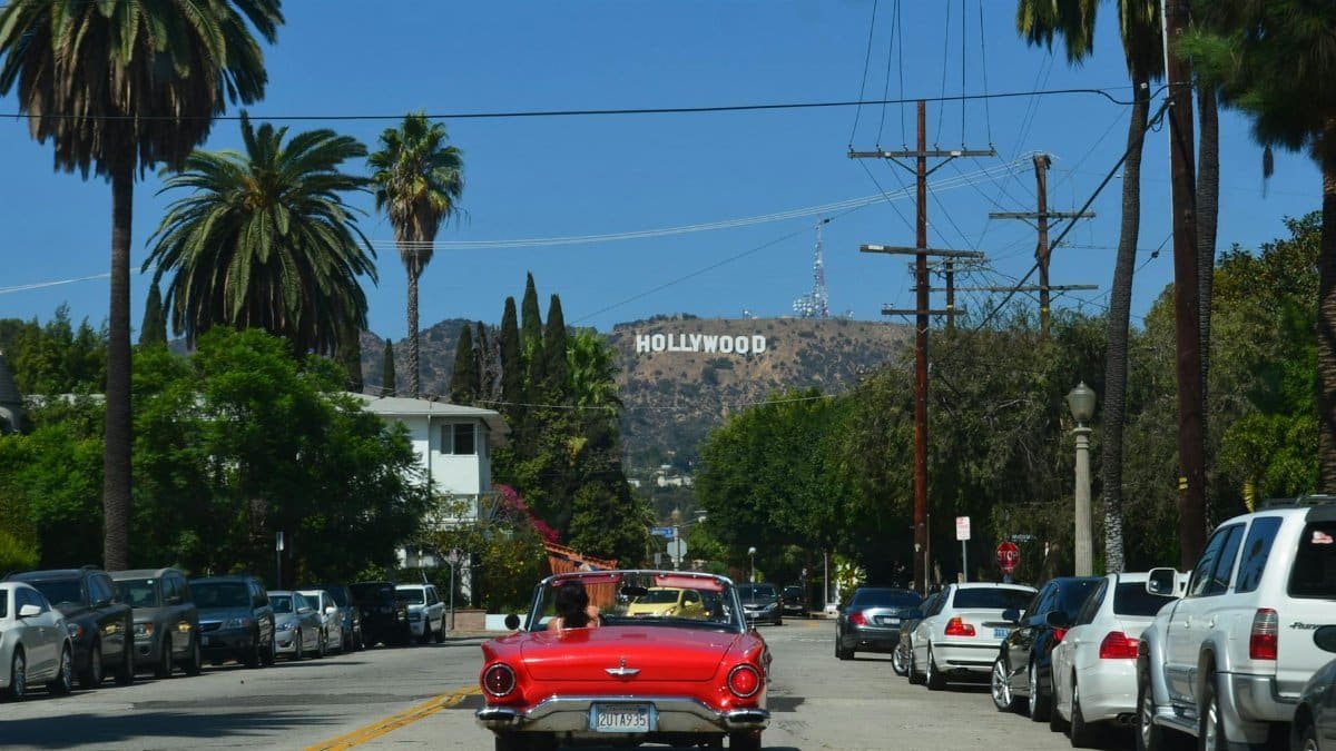 Red convertible driving down a sunny street towards the iconic Hollywood sign in Los Angeles.