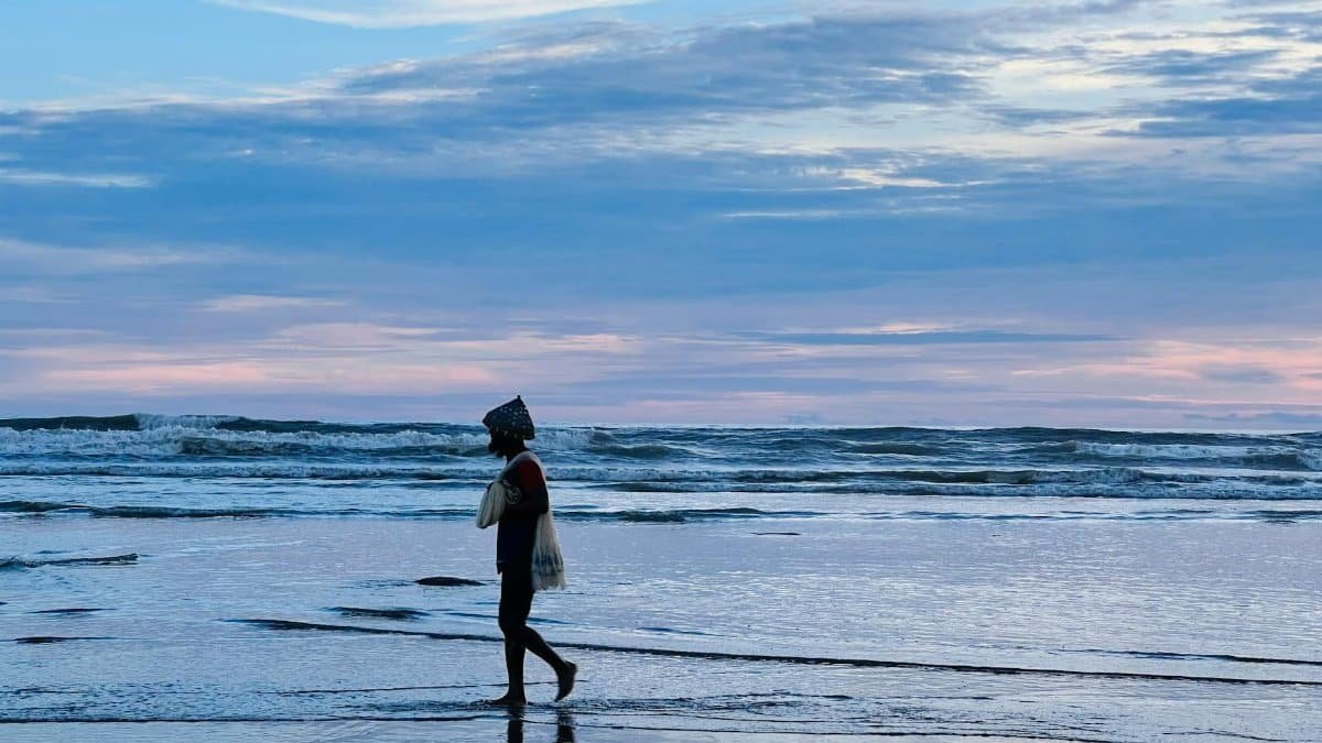 A person walks on Cox's Bazar beach during a peaceful dawn, reflecting tranquil ocean waves and a colorful sky.