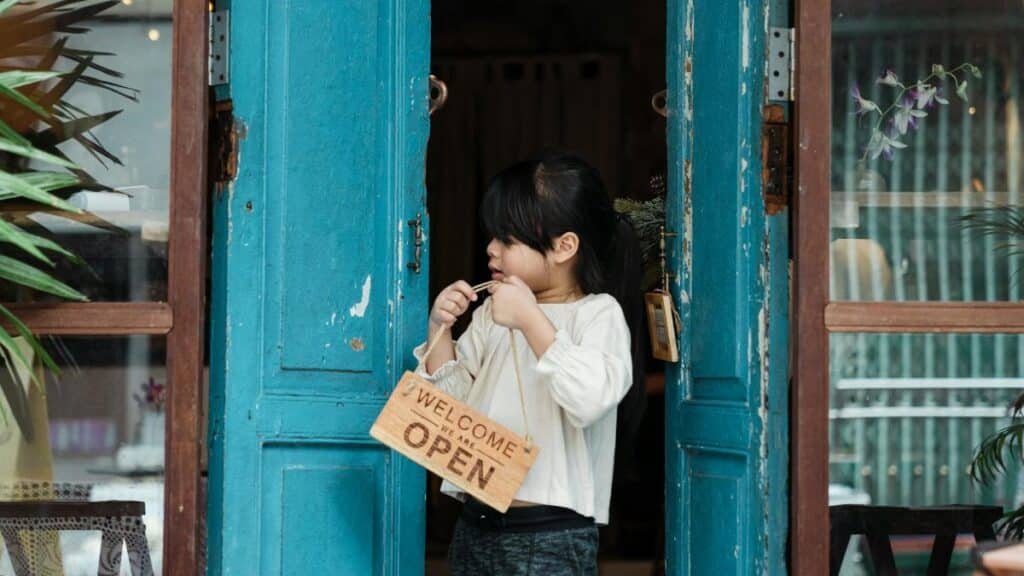 A young girl holding an open welcome sign while standing in a rustic doorway with blue doors.