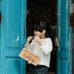 A young girl holding an open welcome sign while standing in a rustic doorway with blue doors.
