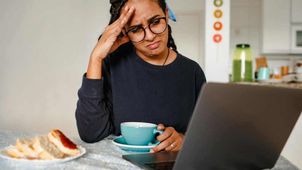 A woman holding her head in frustration while working on a laptop at home, drinking coffee.