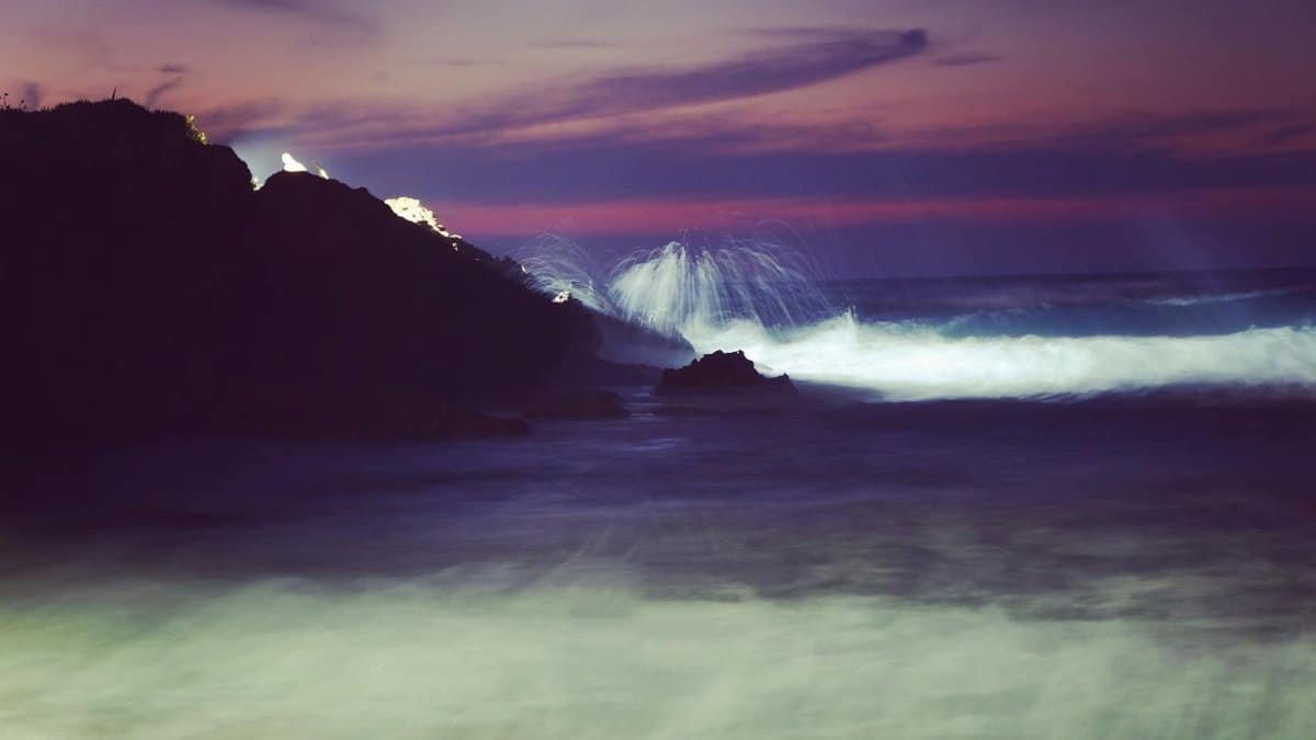Dramatic view of waves crashing on rocks during a colorful sunset in Greece.