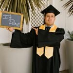 Adult male graduate in cap and gown holding 'I'm Done!' sign outdoors.