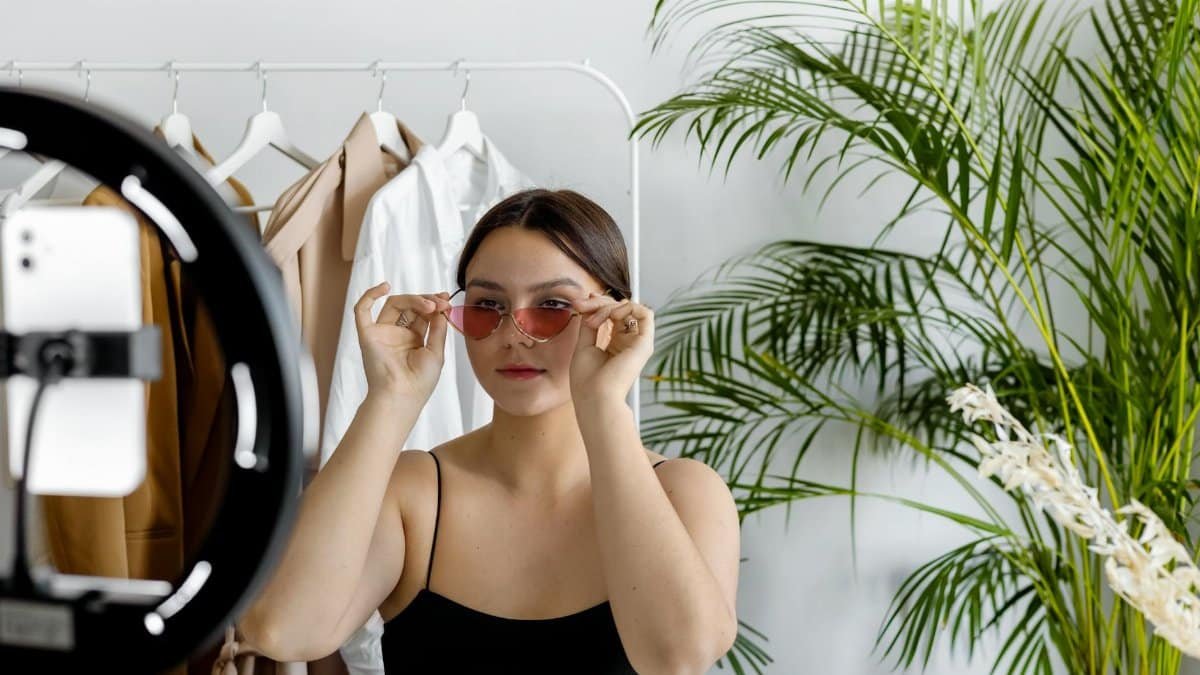 Stylish woman wearing sunglasses recording fashion content with a ring light indoors.