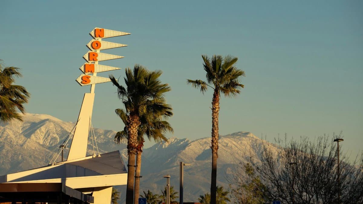 A retro Norms restaurant sign framed by palm trees and snow-capped mountains in Rialto, CA.