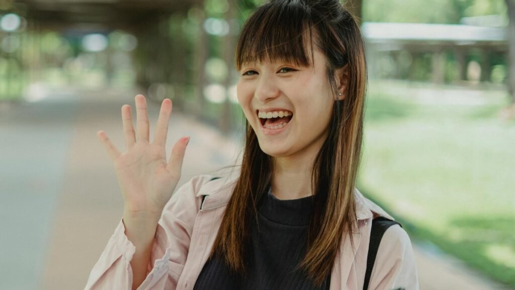 Smiling woman waving hello while standing outdoors on a sunny day.