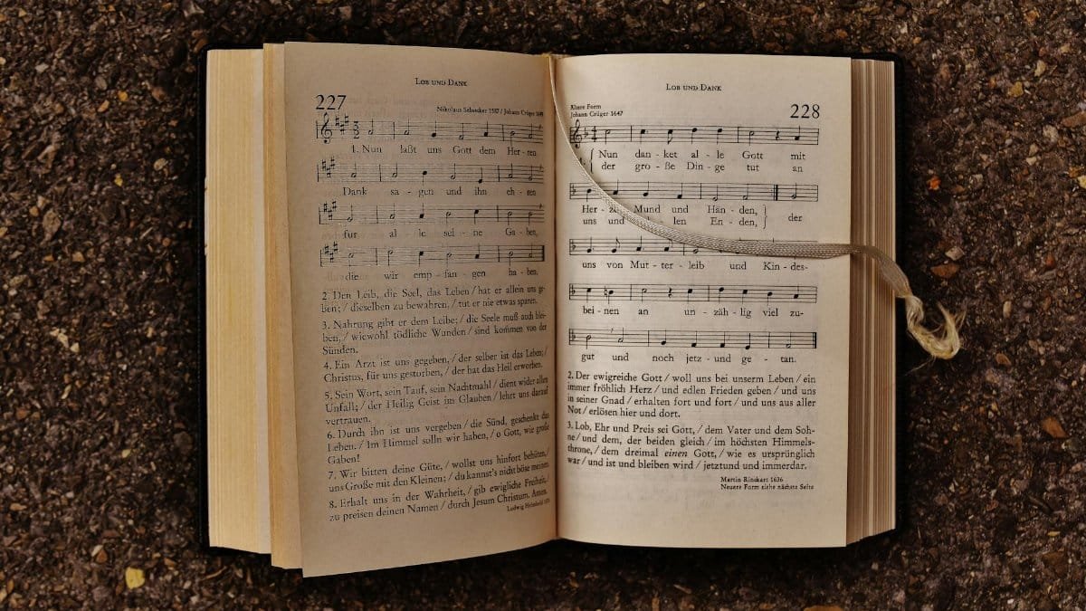 An open hymn book with musical notes laying on earthy ground, featuring sheet music and lyrics.