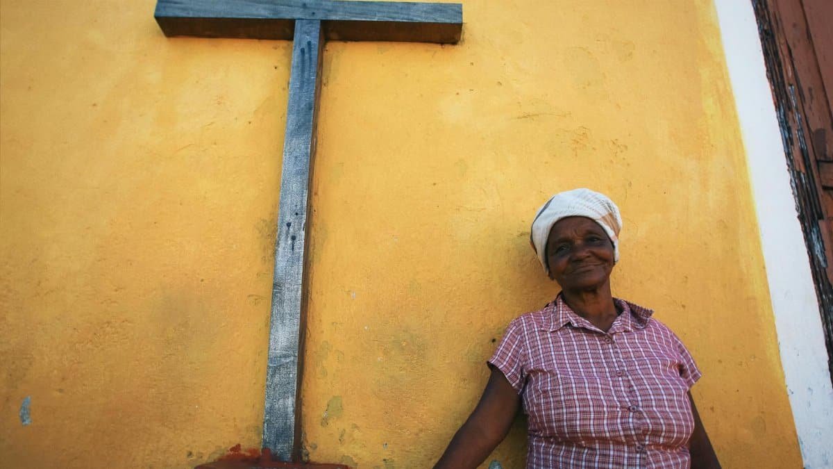 A senior woman stands by a yellow church wall with a cross, reflecting culture and faith.