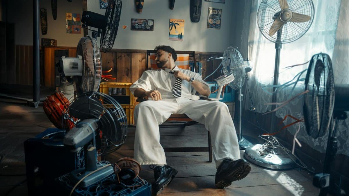 A man in white attire sits surrounded by various fans, cooling off in a warmly lit room.