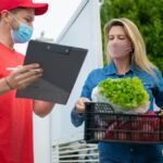 A delivery person in protective gear handing groceries to a customer at home.