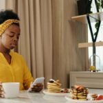 African American woman using smartphone while having pancakes and coffee at a cozy breakfast indoors.