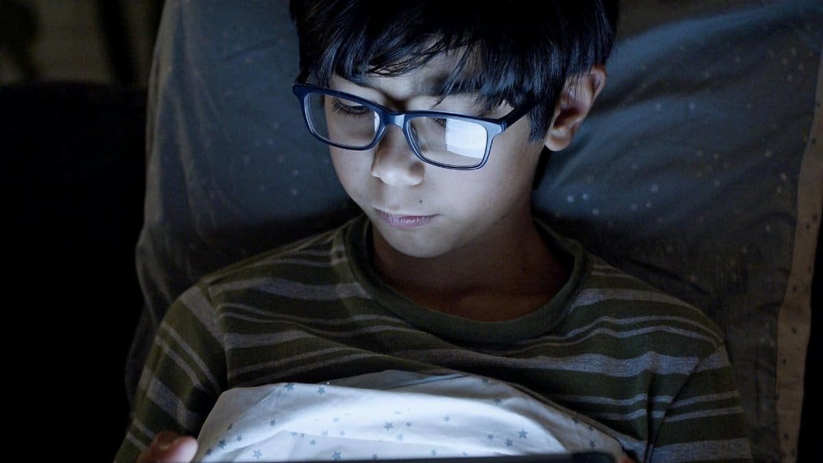 A young boy with glasses uses a digital tablet in bed at night, dimly lit by the screen.