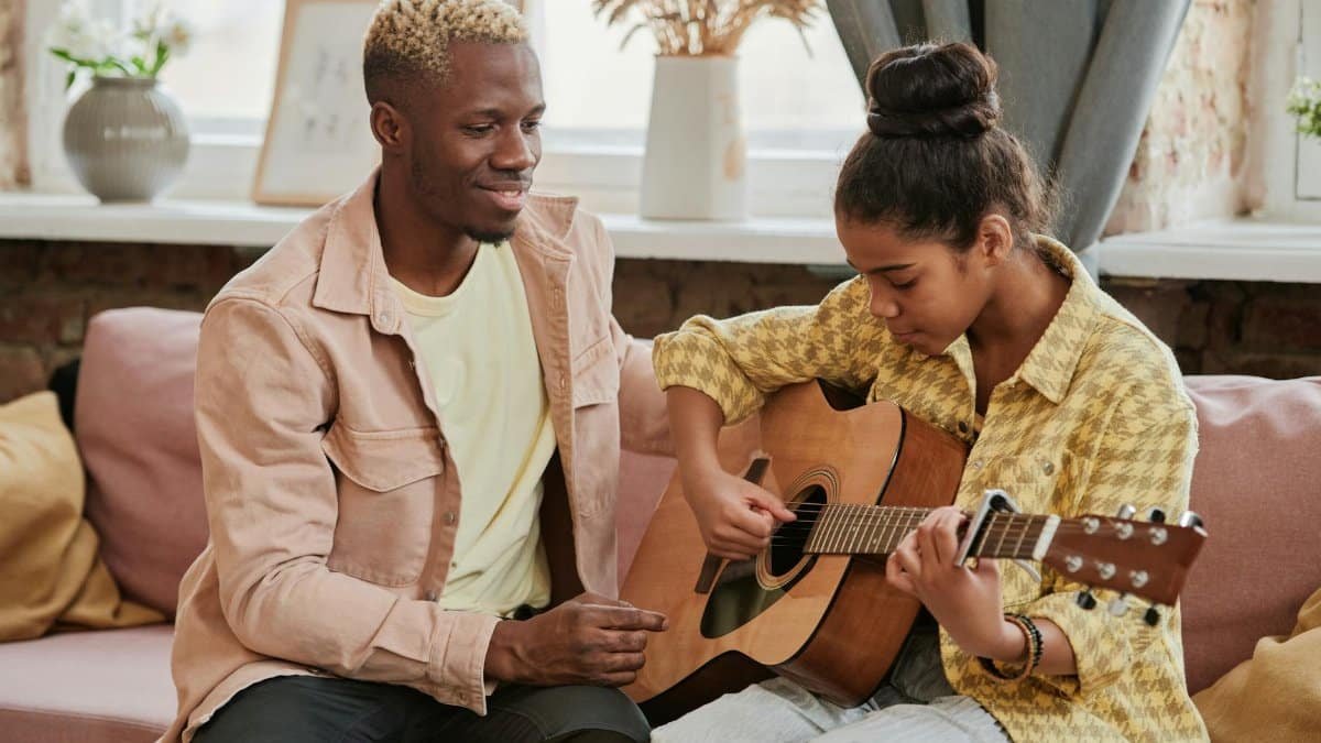 A father teaching his daughter to play acoustic guitar on a cozy indoor couch.