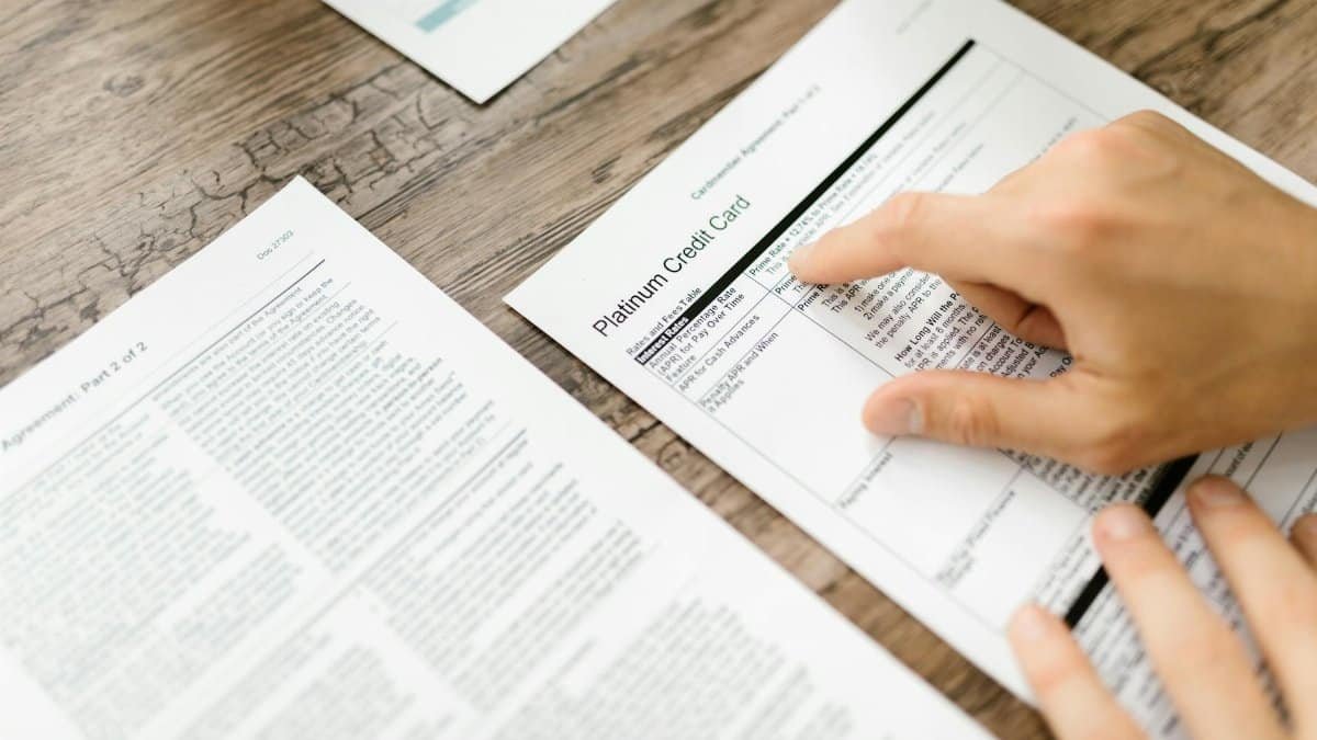 A hand examining a credit card agreement on a wooden desk, highlighting financial review.