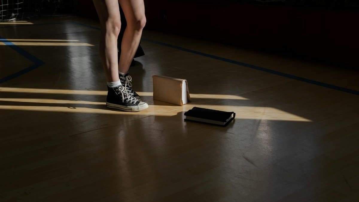 Sunlit scene of student standing in gym with books and sneakers.