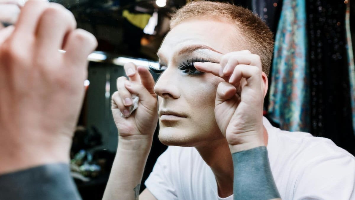 A drag performer applying eyelashes in a mirror before a show, showcasing transformation and artistry.