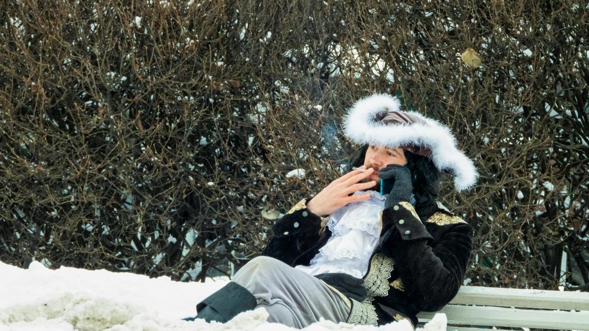 A man in traditional attire relaxes and smokes on a snowy bench, showcasing wintertime outdoor leisure.