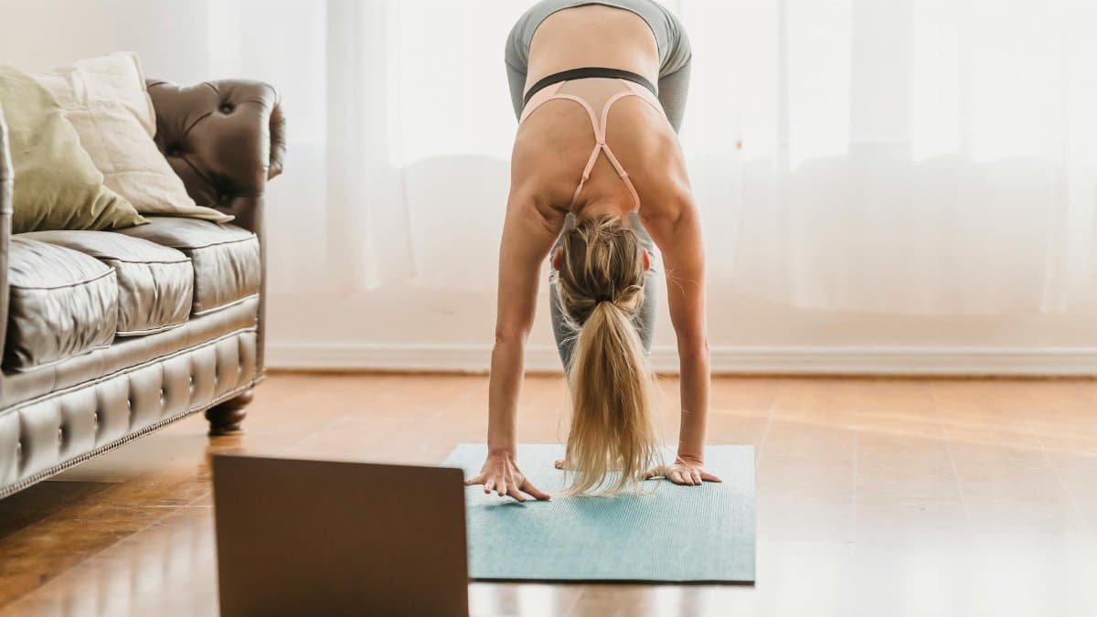 Full body of unrecognizable female in activewear performing standing forward bend asana while standing on mat near laptop during online yoga lesson at home