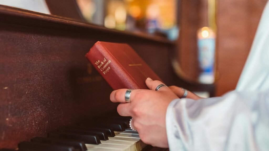 A person holds a hymnal book over piano keys, symbolizing music and faith.