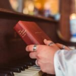 A person holds a hymnal book over piano keys, symbolizing music and faith.
