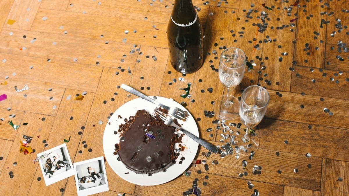 Post-party scene with a chocolate cake, champagne bottle, and confetti on a wooden floor.