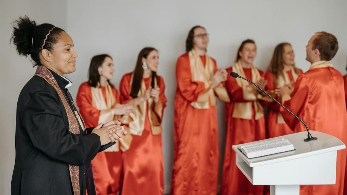 A diverse choir claps and sings passionately in a church setting led by a woman pastor.