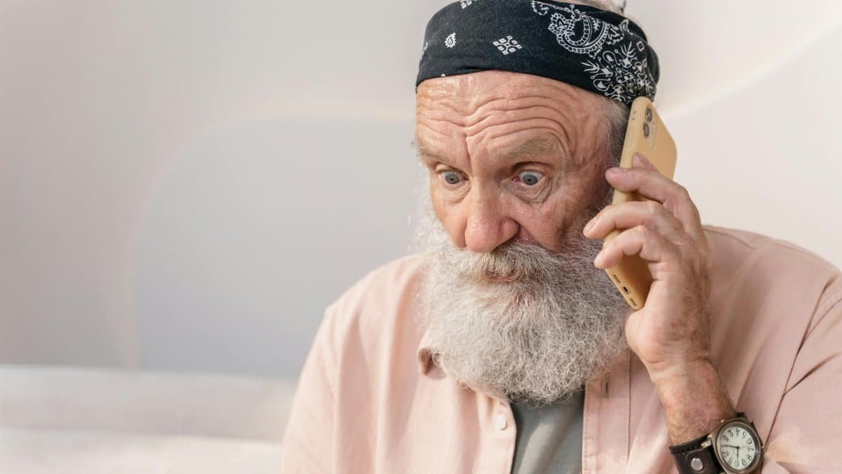 An elderly man with a beard looks surprised while on the phone indoors.