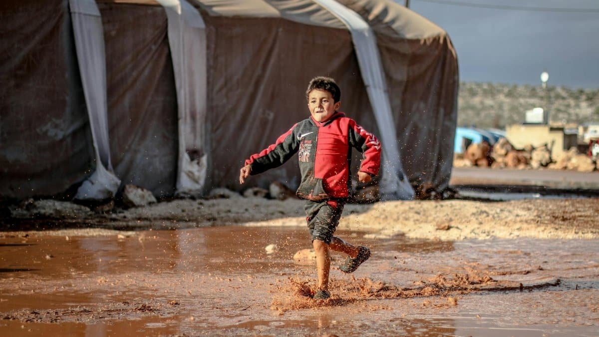 A joyful young boy runs through a muddy puddle in a refugee camp in Idlib, Syria, encapsulating resilience and innocence.