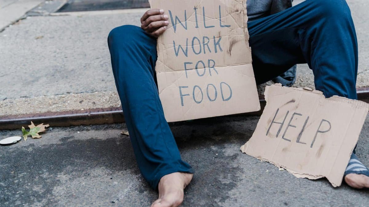 A homeless person sitting on the street holding a cardboard sign with the message 'Will work for food'.