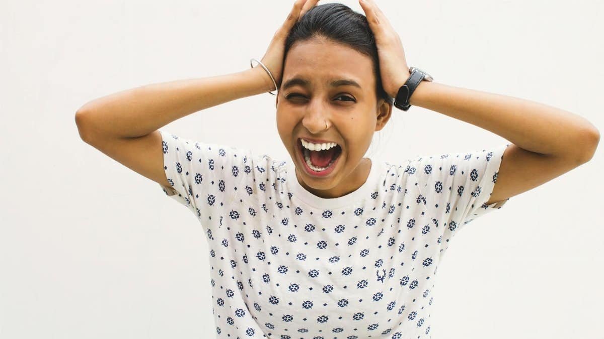 A young woman in a casual shirt screams with hands on her head against a white background.