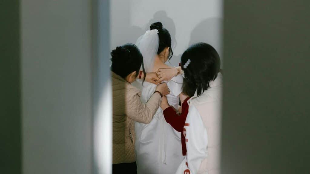 A bride getting ready with help from others, captured from behind in a bridal room.