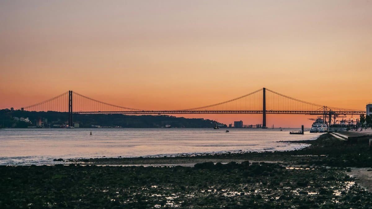 A breathtaking sunset over Lisbon's 25 de Abril Bridge, highlighting its reflection on the water.