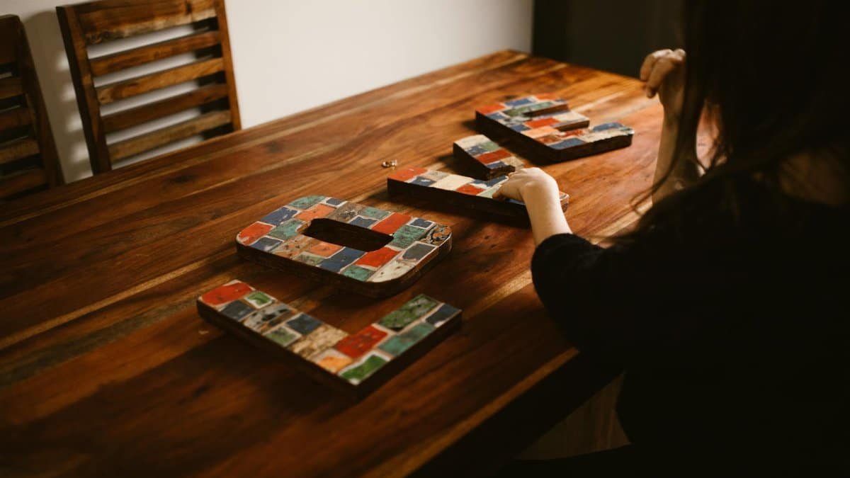 A woman arranging colorful mosaic letters on a wooden table indoors, creating art.