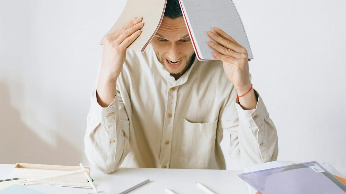 A stressed adult male worker sits at a desk with open notebooks, exhibiting signs of frustration and burnout.