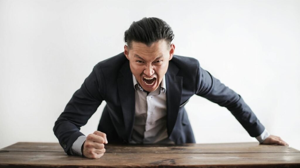 Expressive angry businessman in formal suit looking at camera and screaming with madness while hitting desk with fist