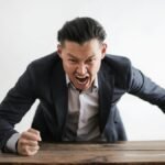Expressive angry businessman in formal suit looking at camera and screaming with madness while hitting desk with fist