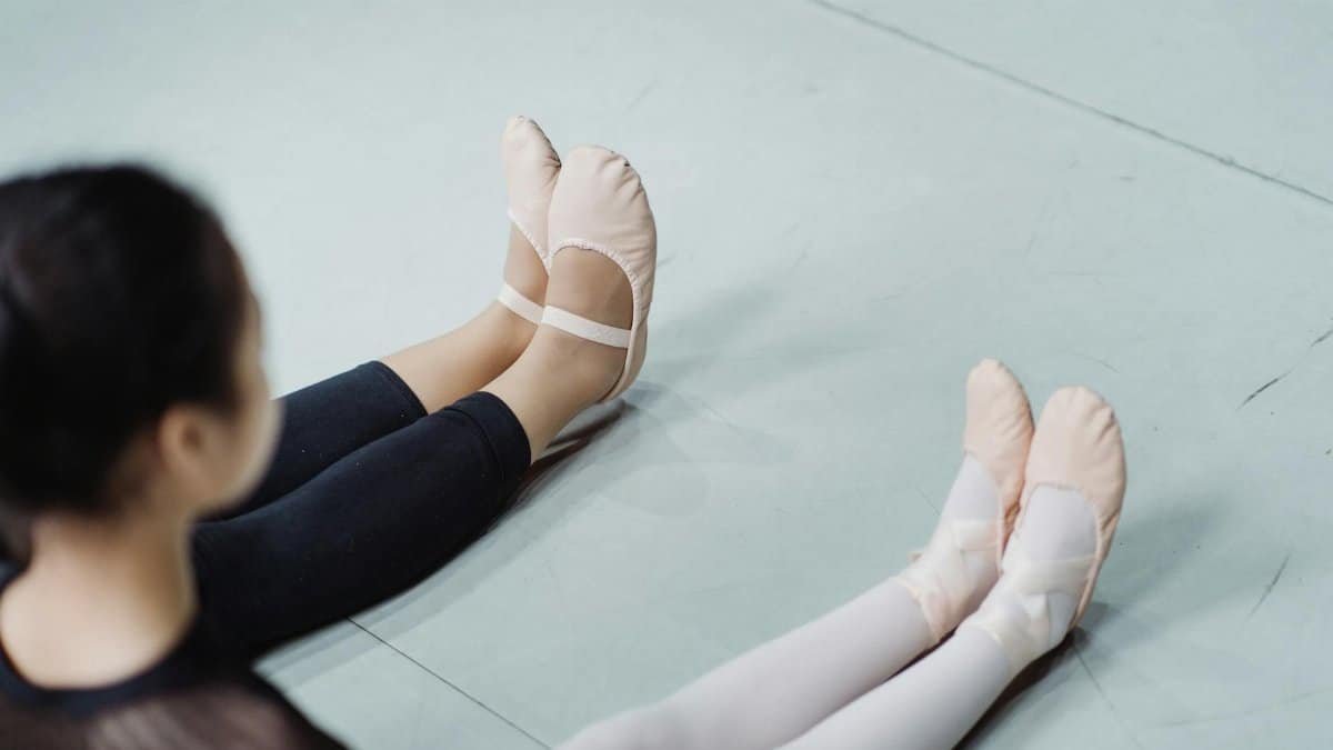 Two ballet dancers sitting on the floor practicing with focus on their feet and slippers in a studio setting.