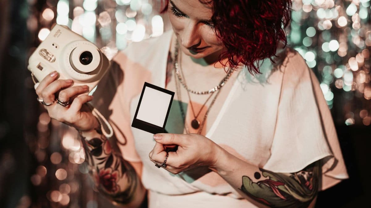 Tattooed woman with red hair holding an instant camera and a blank photo indoors at a party.