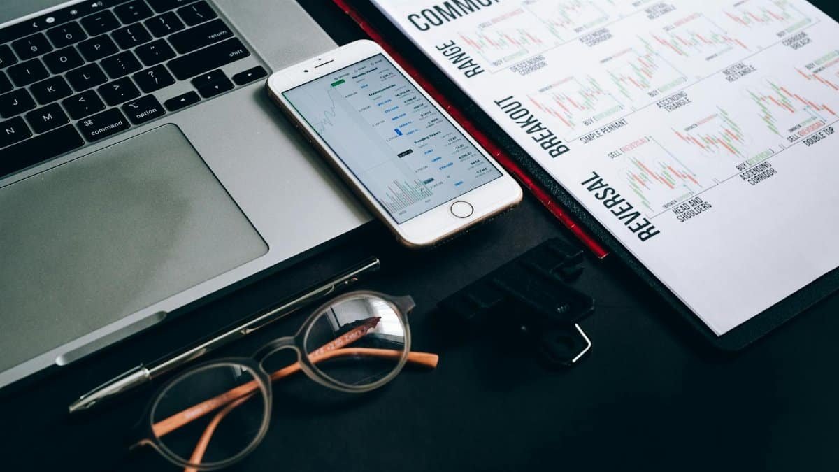 Workstation with laptop, smartphone, eyeglasses, financial charts, and pen for trading analysis.