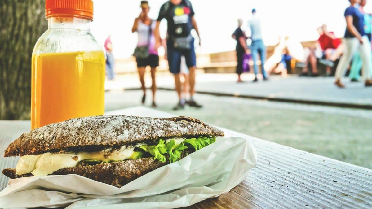 Close-up of a sandwich and orange juice on a table with people in the park.
