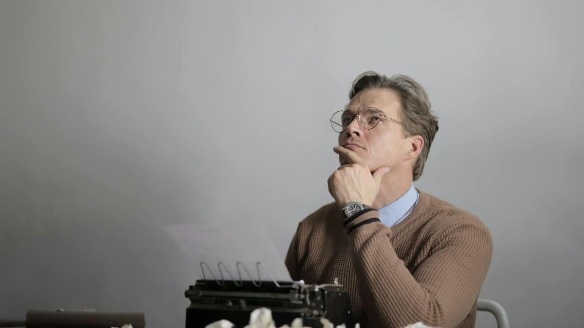 Man thinking deeply with a typewriter, surrounded by crumpled paper, in an indoor setting.
