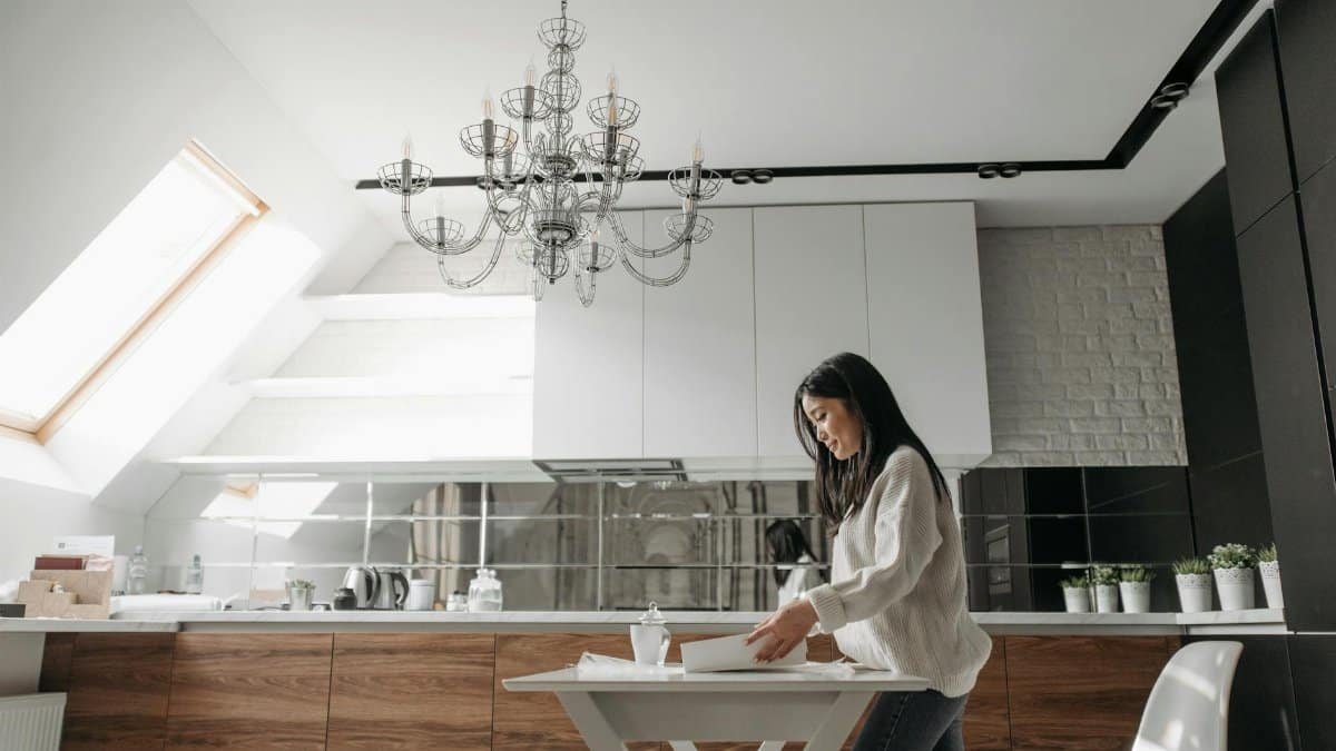 Asian woman unpacking in a modern kitchen with wooden cabinets and chandelier.