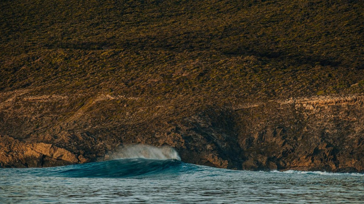 A large ocean wave crashes against rugged cliffs with tranquil blue water, showcasing nature's power and beauty.