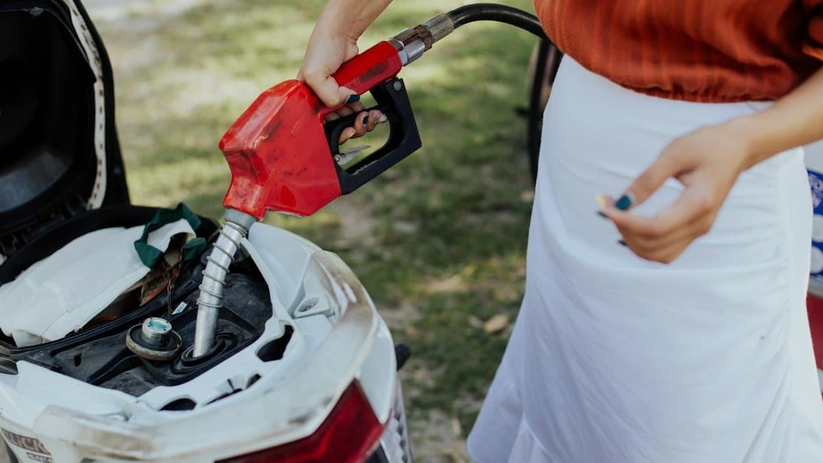 A person refuels a scooter with gasoline using a red nozzle, outdoors on a sunny day.