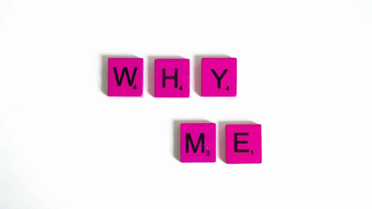 Pink Scrabble tiles form the words 'Why Me' on a white background, emphasizing questioning and introspection.