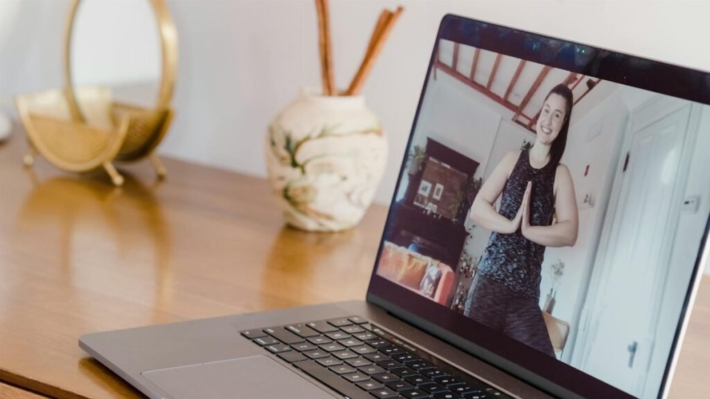 A woman participates in an online yoga class, seen on a laptop screen at home.