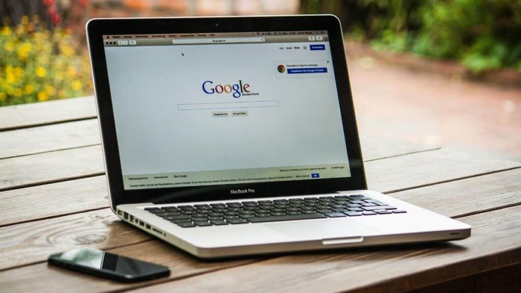 A MacBook Pro displaying Google Search on a wooden table outdoors, next to a smartphone.
