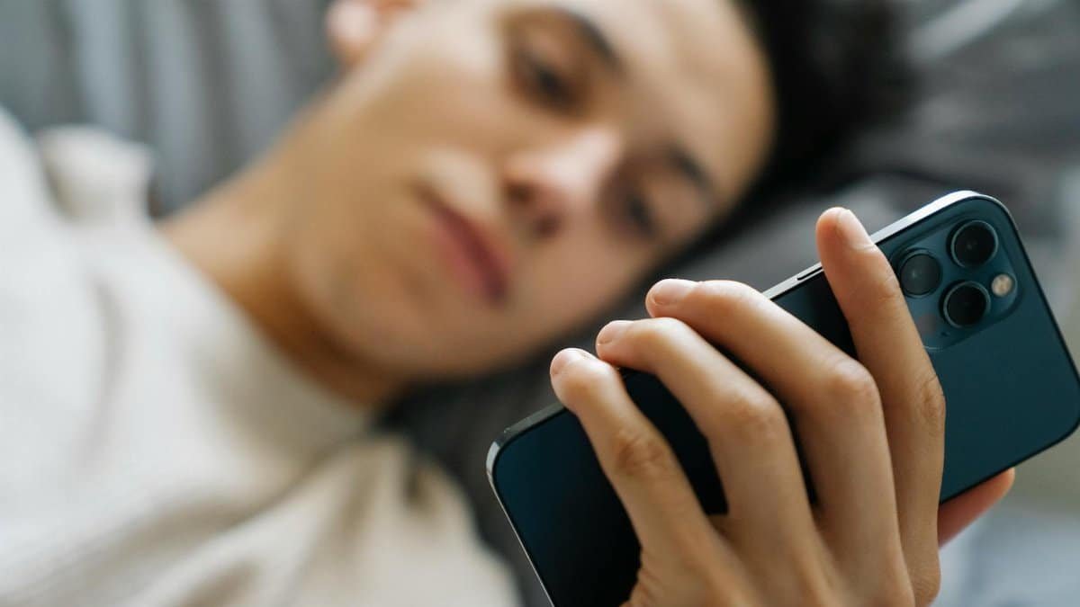 Soft focus of young guy lying on bed and using modern smartphone in weekend morning at home